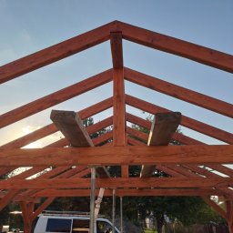 IVAN SHEVCHUK PRO100BUD - Konstrukcja drewniana dachu w trakcie budowy, widoczne belki w kolorze brązowym, wsporniki metalowe i biały samochód dostawczy w tle.