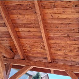 IVAN SHEVCHUK PRO100BUD - Drewniana konstrukcja z belek i desek pomalowanych bejcą na ciepły, brązowy kolor, widok z dołu na zadaszenie, z fragmentami budynków i nieba w tle.