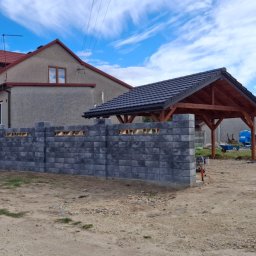 IVAN SHEVCHUK PRO100BUD - Nowoczesny, szary, bloczkowy mur ogrodzeniowy z otworami wentylacyjnymi w trakcie budowy, z widocznym domem i drewnianą wiatą w tle, na piaszczystym podłożu.