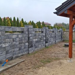 IVAN SHEVCHUK PRO100BUD - Wznoszenie ogrodzenia z szarej kostki betonowej, widoczne rurki instalacyjne, w tle drewniana altana i tuje