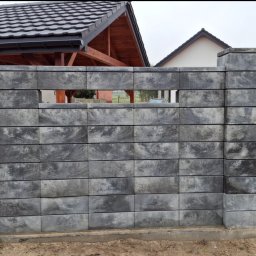 IVAN SHEVCHUK PRO100BUD - Szary, betonowy płot modułowy z pustakami o nieregularnym wzorze, z widocznym prześwitem i fragmentem zabudowań w tle oraz białym vanem po prawej stronie na żwirowej drodze.