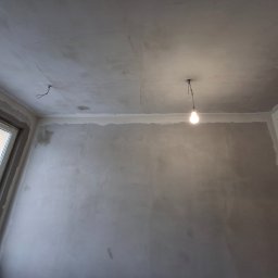 IVAN SHEVCHUK PRO100BUD - Surowa ściana w pokoju po nałożeniu gładzi szpachlowej, widoczne niedociągnięcia i wystający kabel z sufitu oświetlony wiszącą żarówką, okno po lewej stronie.