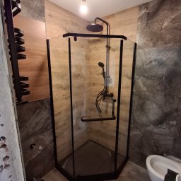 IVAN SHEVCHUK PRO100BUD - Narożna kabina prysznicowa z czarnymi profilami i deszczownicą, obok grzejnik drabinkowy w trakcie montażu, widoczne fragmenty ściany z uszkodzonym tynkiem i wiadro z zaprawą.