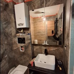 IVAN SHEVCHUK PRO100BUD - Niedokończona łazienka z marmurowymi płytkami, wiszącą toaletą, białą umywalką na czarnej szafce i lustrem z widocznym odbiciem osoby robiącej zdjęcie, z zamontowanym piecem gazowym...