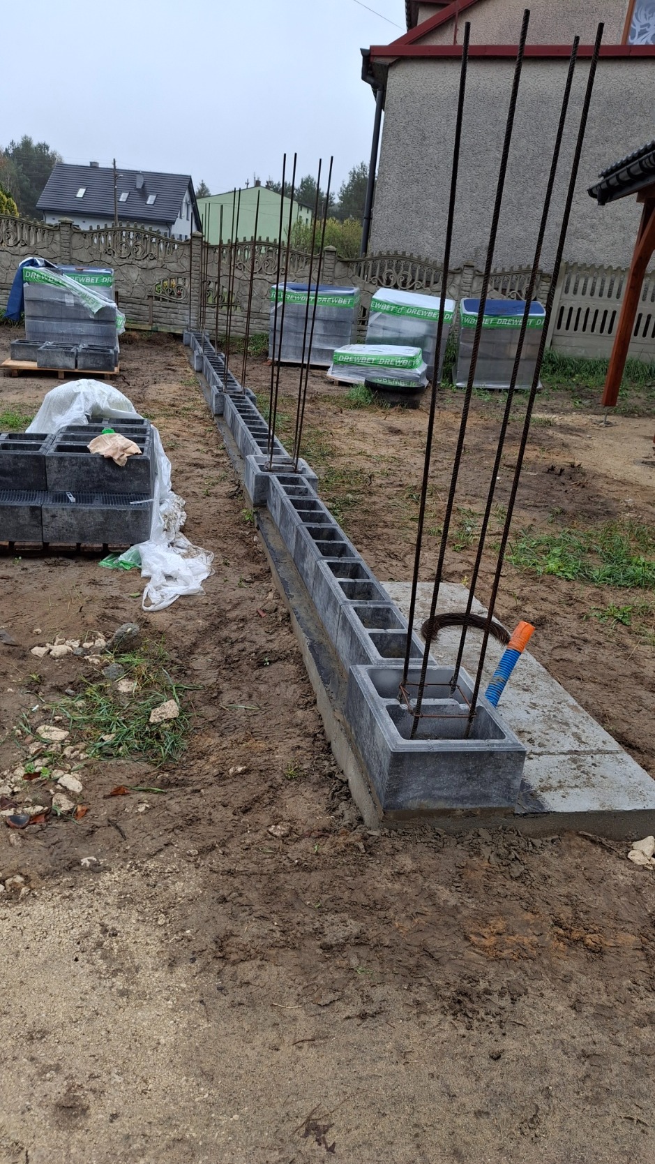 Budowa fundamentu ogrodzenia z betonowych bloczków z wystającymi prętami zbrojeniowymi, na grząskim terenie, w tle domy mieszkalne.