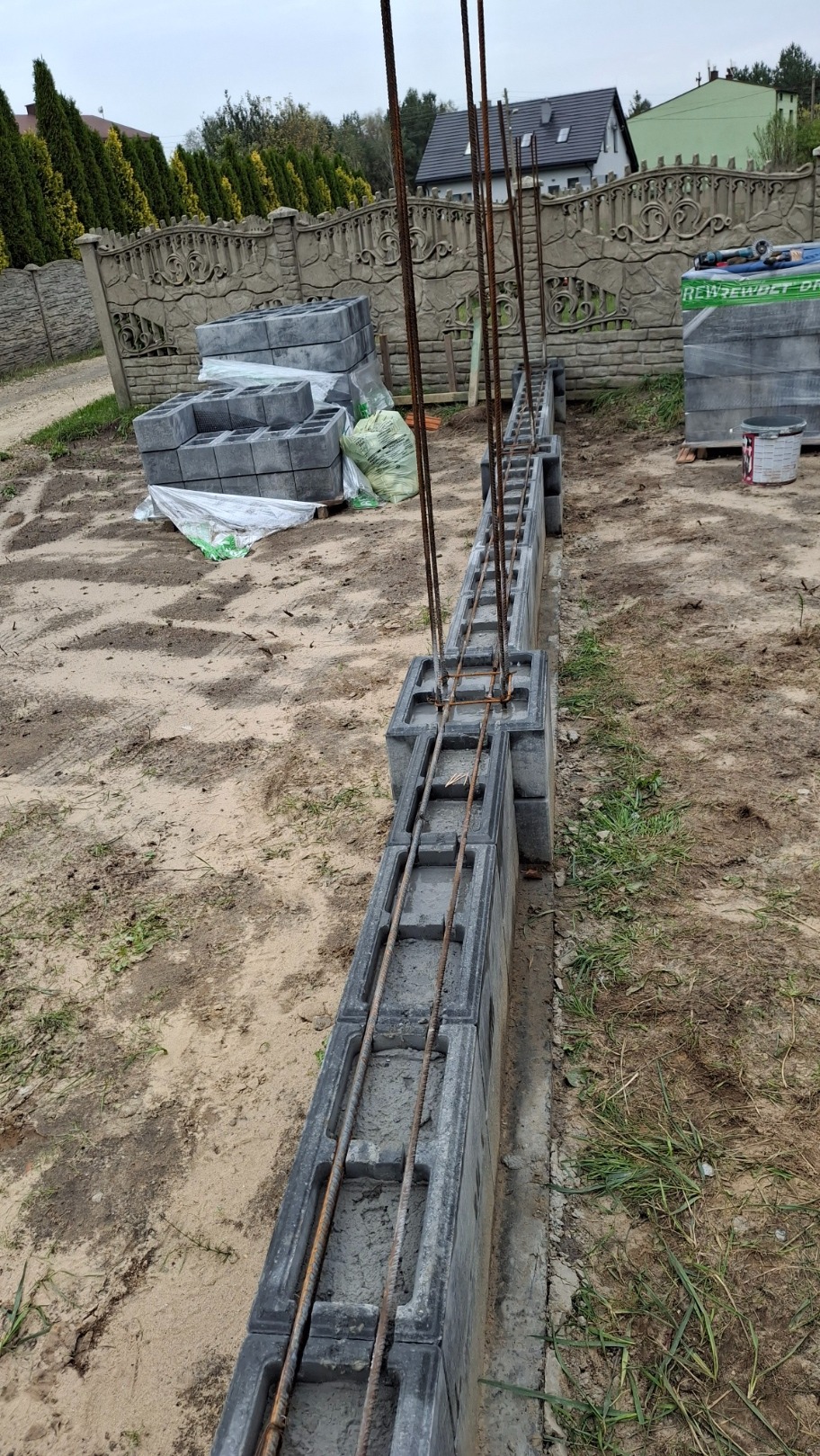 Budowa fundamentu ogrodzenia z szarych bloczków betonowych zbrojonych stalowymi prętami, widoczne ułożone bloczki i materiały budowlane na piaszczystym podłożu, w tle ozdobny, betonowy płot i dom.