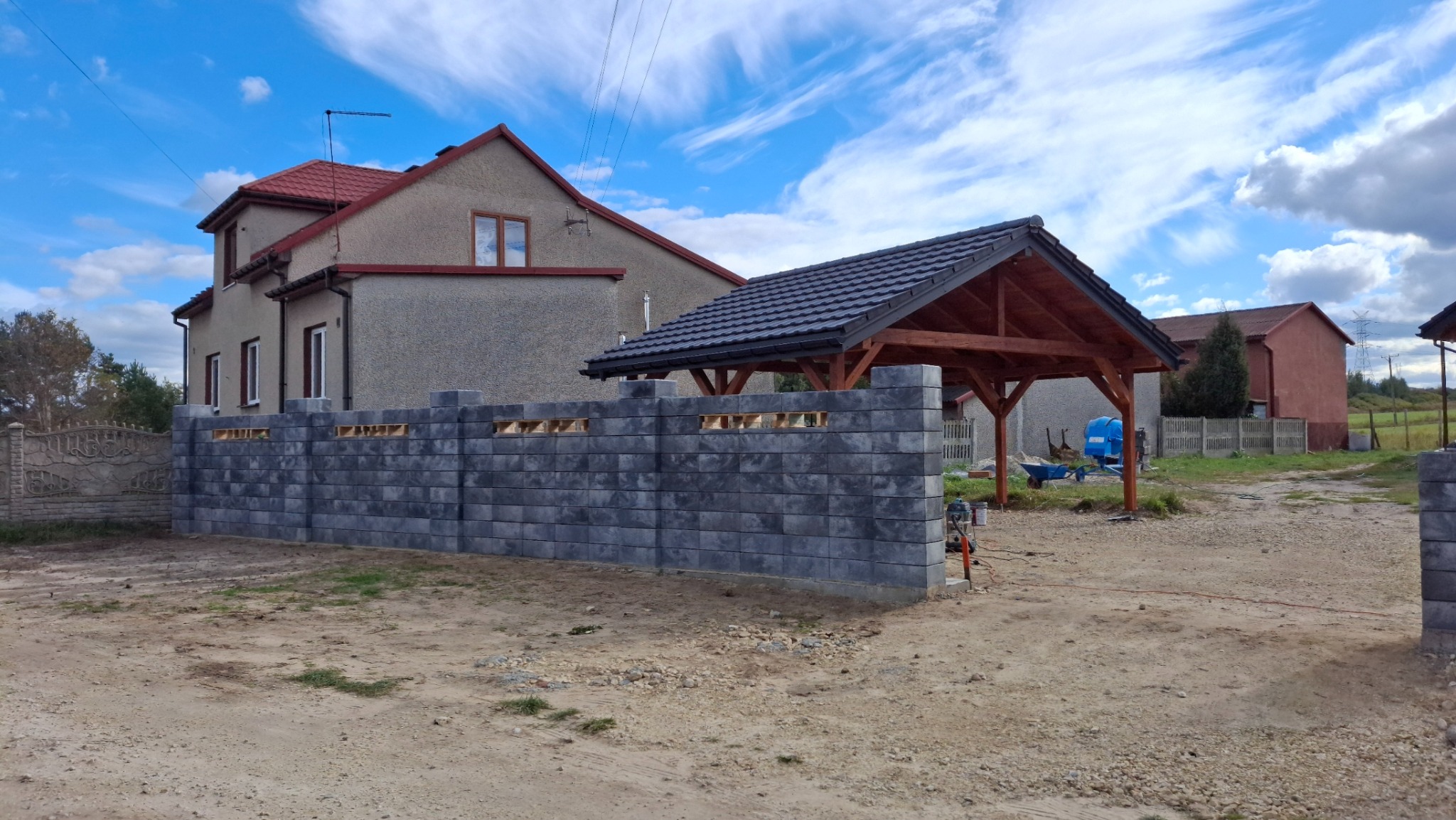 Nowoczesny, szary, bloczkowy mur ogrodzeniowy z otworami wentylacyjnymi w trakcie budowy, z widocznym domem i drewnianą wiatą w tle, na piaszczystym podłożu.