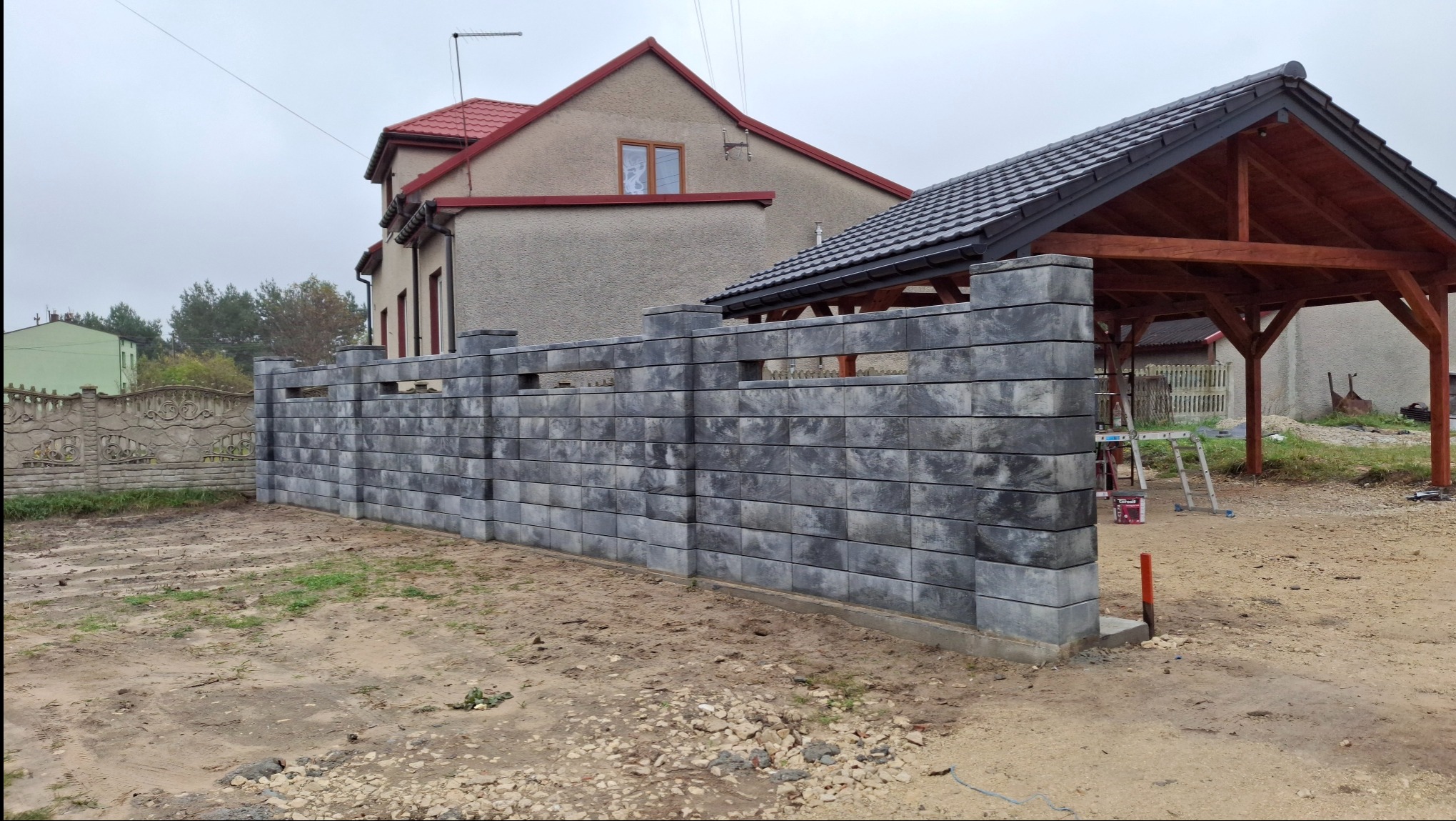 Nowoczesny, szary ogrodzenie modułowe z pustaków betonowych, z widocznymi przerwami między elementami, na tle domu z czerwoną dachówką i drewnianej wiaty.