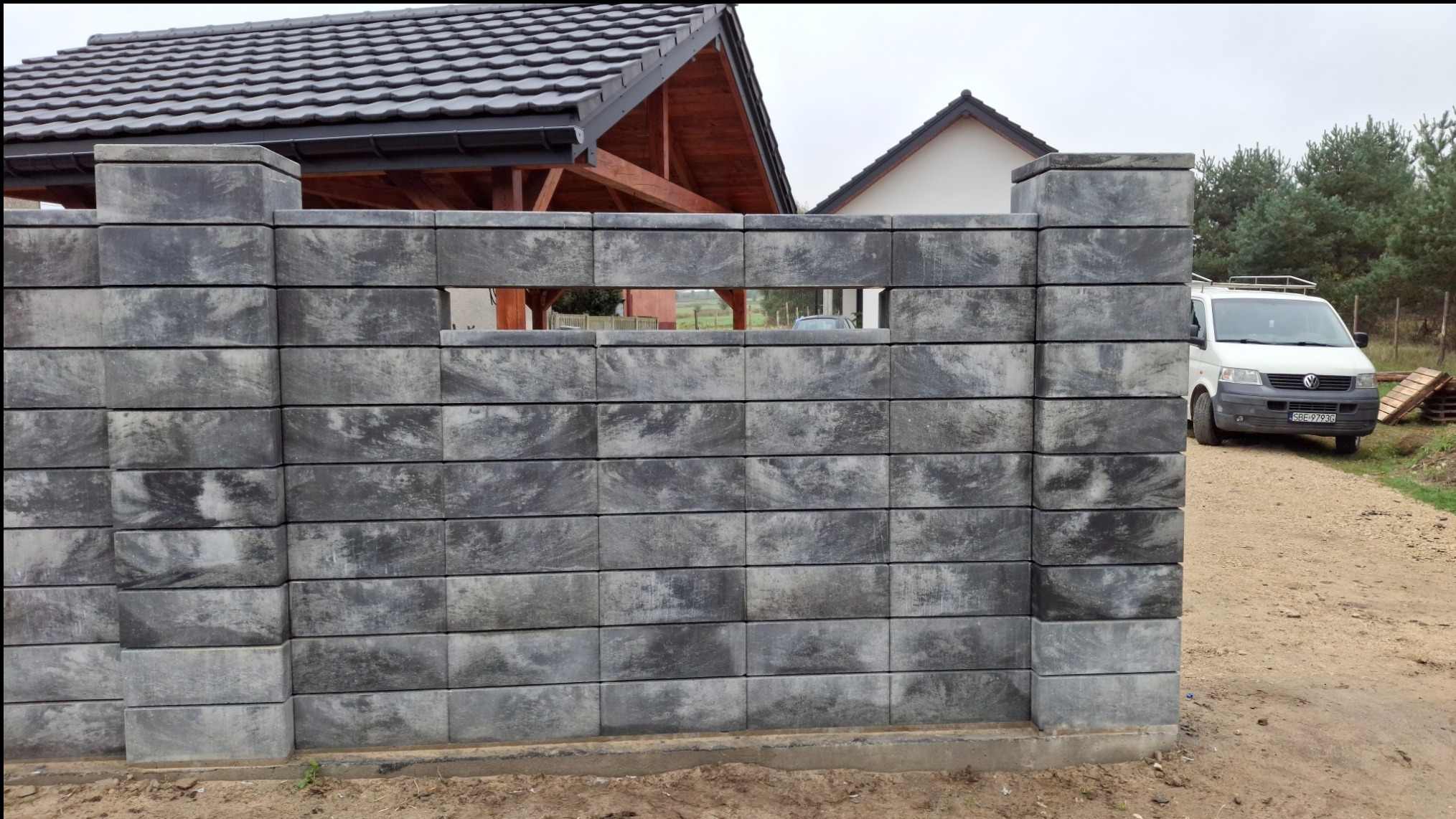 Szary, betonowy płot modułowy z pustakami o nieregularnym wzorze, z widocznym prześwitem i fragmentem zabudowań w tle oraz białym vanem po prawej stronie na żwirowej drodze.