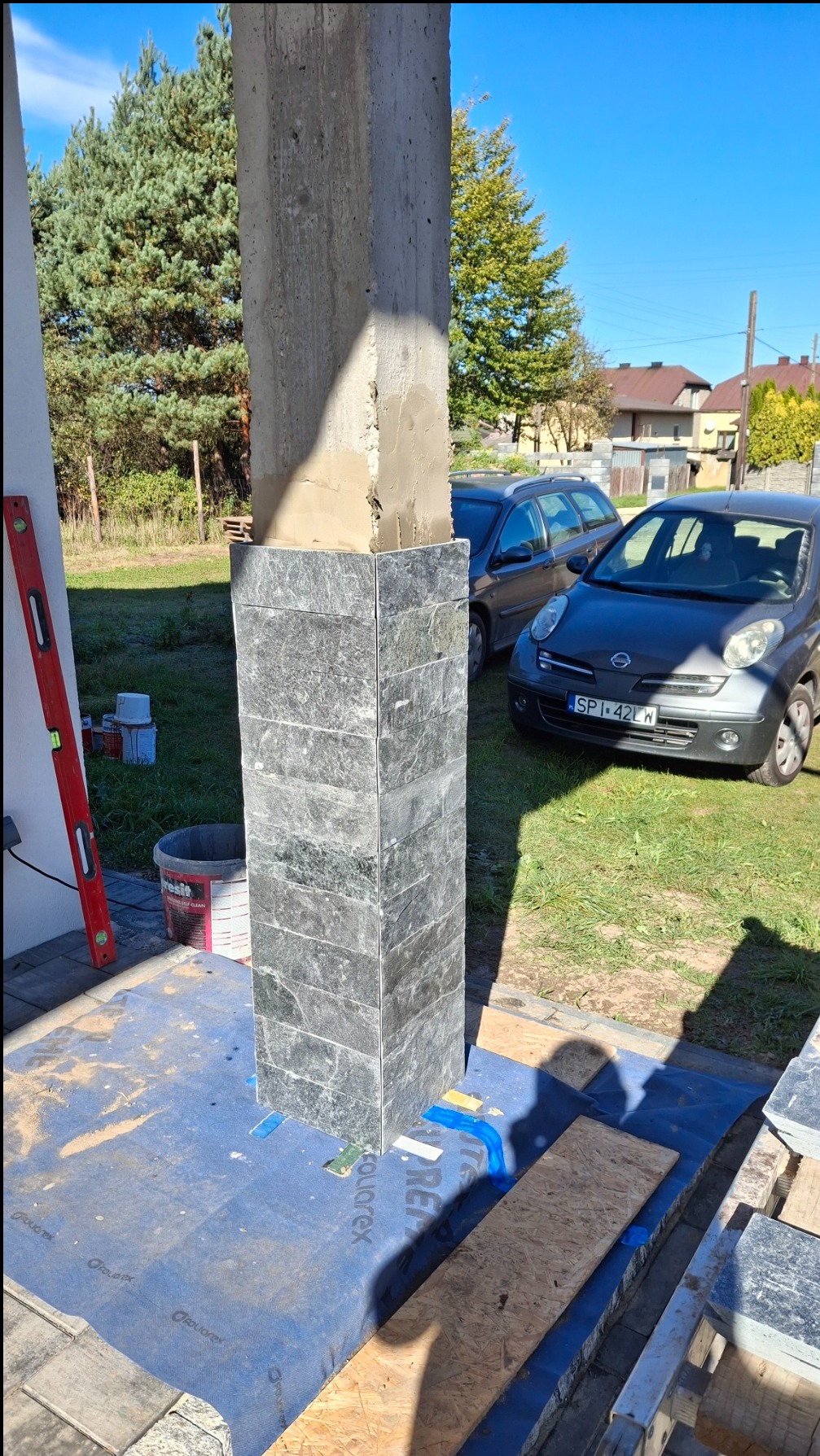 Okleina z kamienia naturalnego na betonowym słupie, częściowo ukończona, widoczne narzędzia i materiały budowlane, samochody w tle.