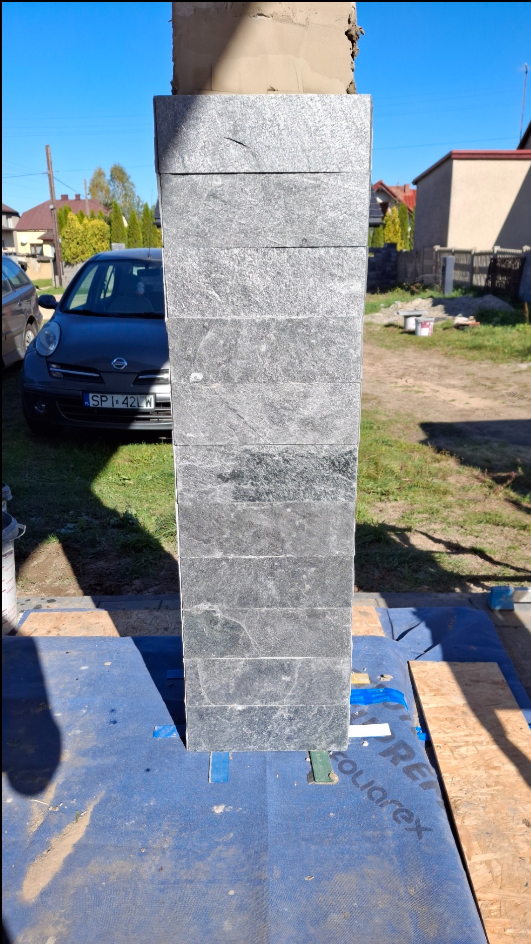 Fragment słupa ogrodzeniowego w trakcie obkładania szarymi płytkami imitującymi kamień, widoczne zabezpieczenie podłoża folią budowlaną i zaparkowany samochód w tle.
