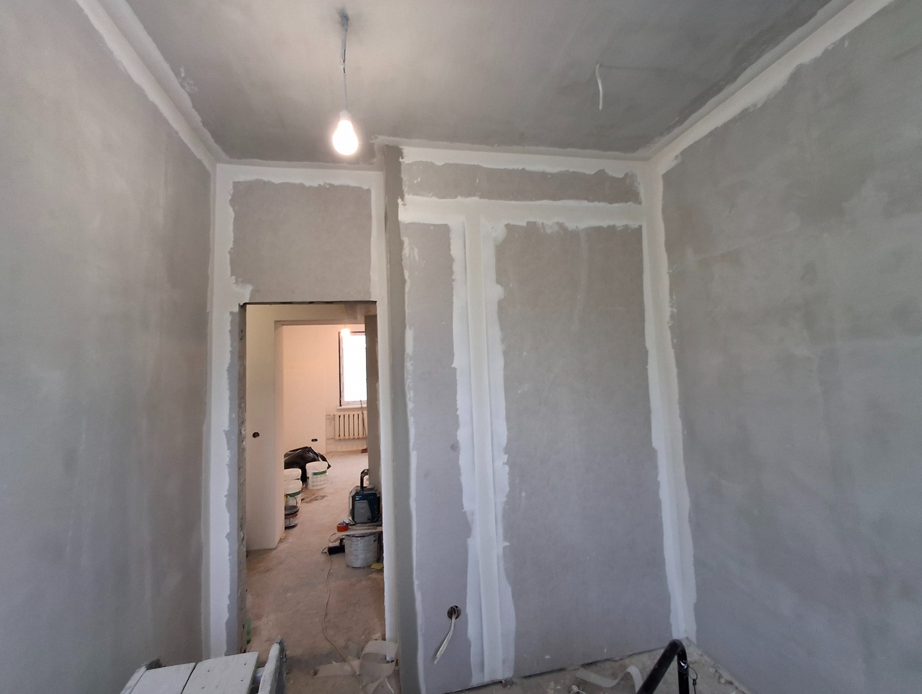 Ściany w surowym stanie podczas szpachlowania, widoczne puszki z farbą w tle, sufit z wiszącą żarówką, wstępny etap prac.