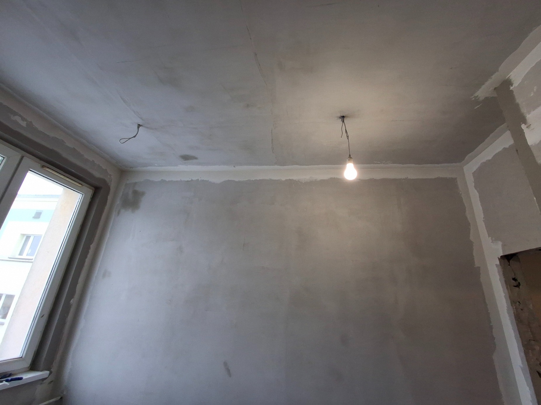 Surowa ściana w pokoju po nałożeniu gładzi szpachlowej, widoczne niedociągnięcia i wystający kabel z sufitu oświetlony wiszącą żarówką, okno po lewej stronie.