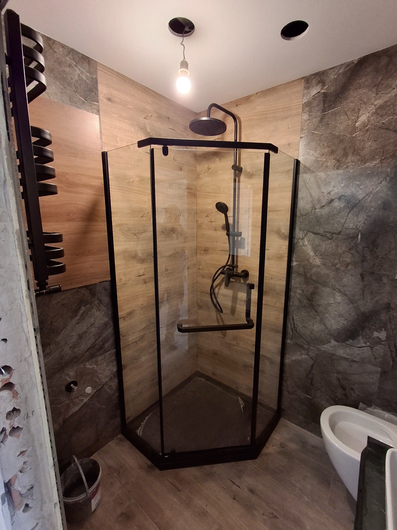 Narożna kabina prysznicowa z czarnymi profilami i deszczownicą, obok grzejnik drabinkowy w trakcie montażu, widoczne fragmenty ściany z uszkodzonym tynkiem i wiadro z zaprawą.