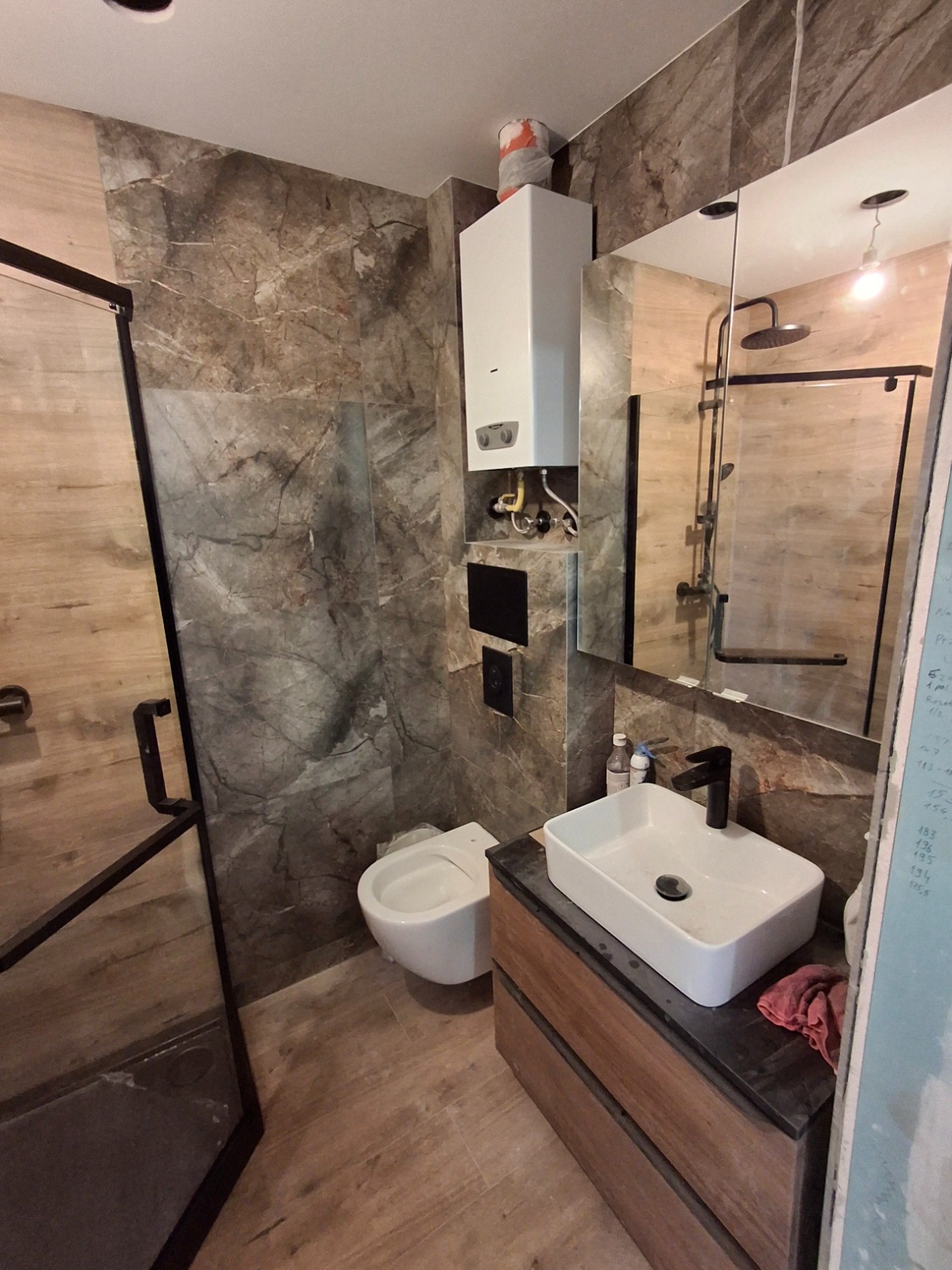 Nowoczesna łazienka w odcieniach brązu i szarości z marmurkowymi płytkami, wiszącą toaletą, szafką z umywalką w stylu industrialnym i szklaną kabiną prysznicową z czarnymi ramami.