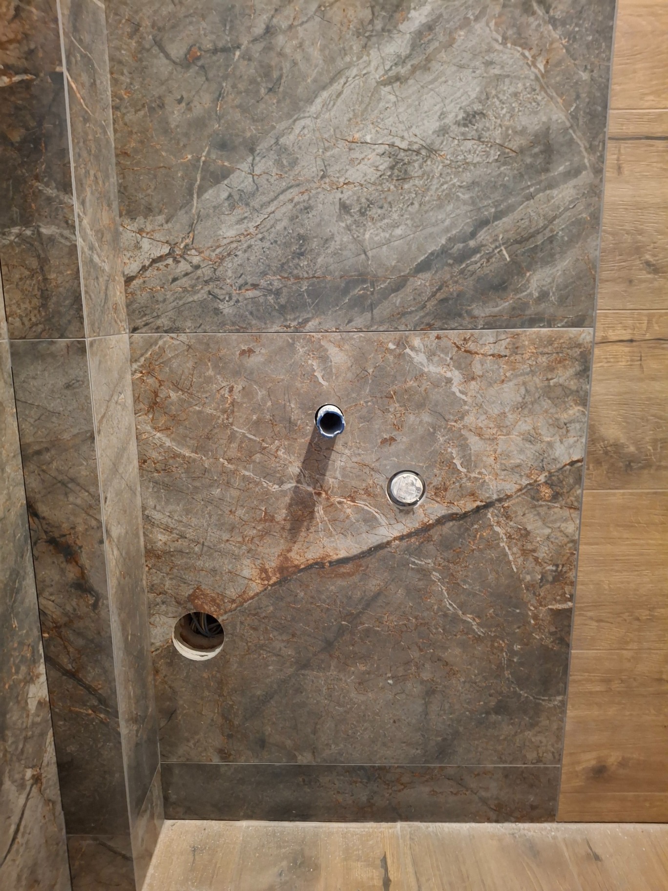 Fragment ściany wyłożonej płytkami imitującymi marmur w odcieniach szarości i brązu, z widocznymi otworami na instalacje wodno-kanalizacyjne i elektryczne, sąsiadująca z panelami imitującymi drewno.