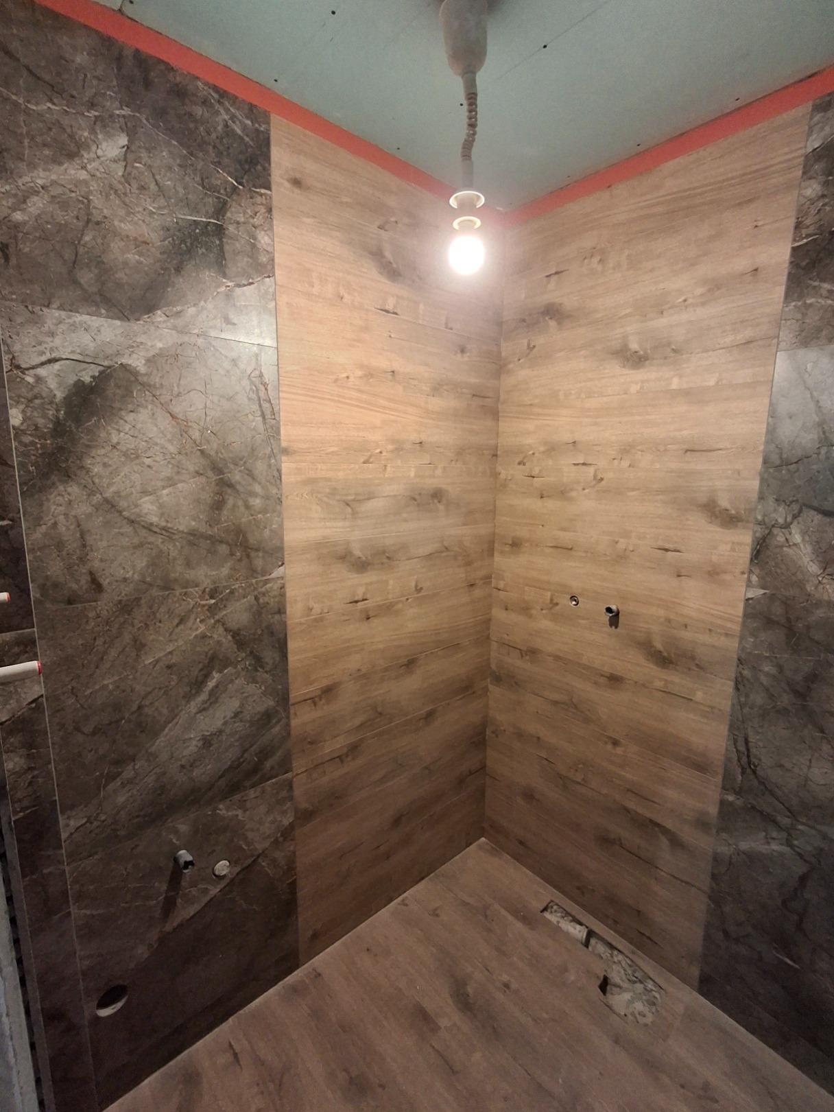 Wnętrze łazienki w trakcie prac: ściany wyłożone częściowo marmurowymi płytkami, częściowo panelami imitującymi drewno, surowy sufit z wystającą żarówką, widoczne instalacje hydrauliczne.