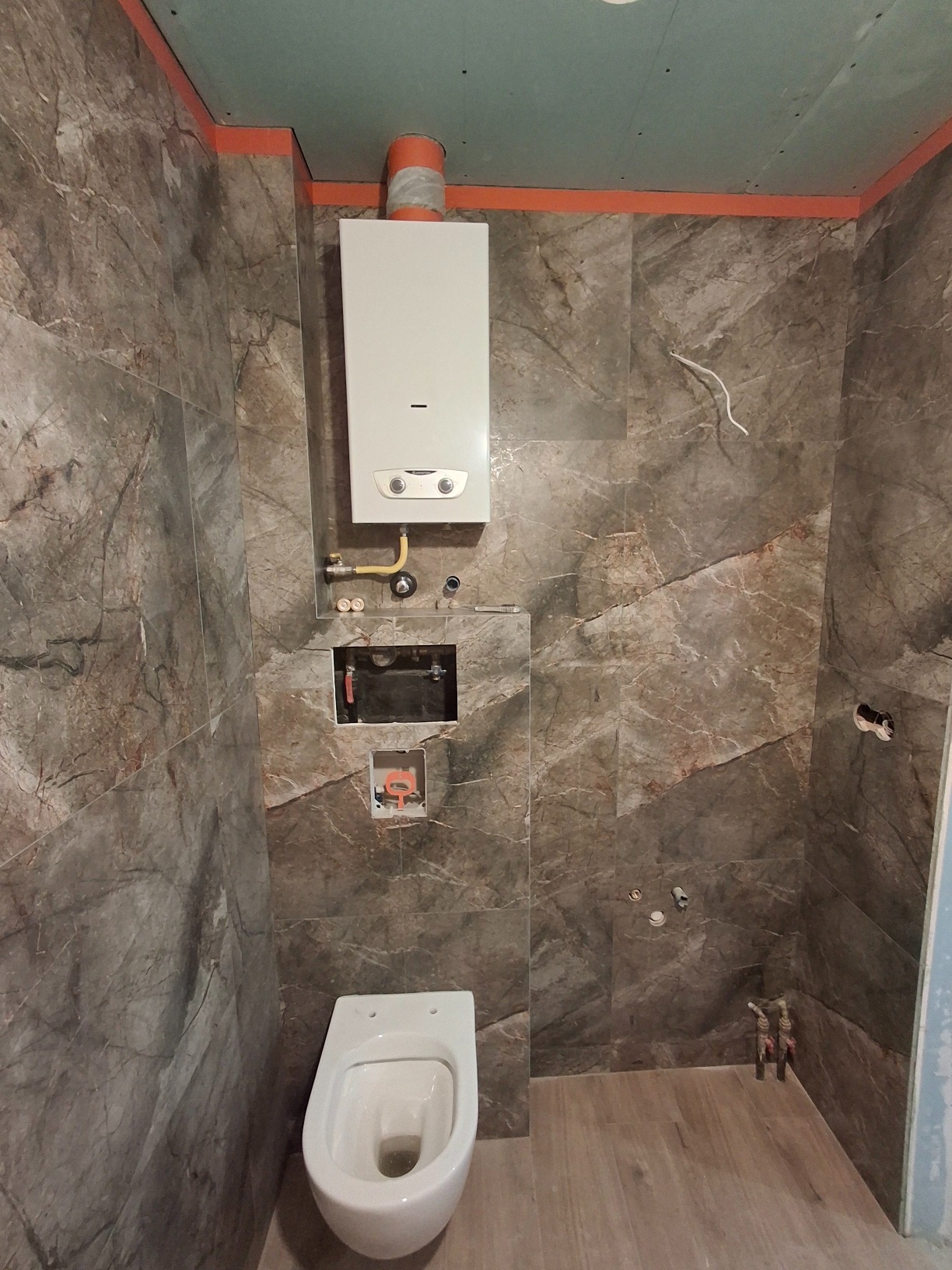 Wnętrze łazienki w trakcie prac: widoczne marmurowe płytki na ścianach, częściowo zamontowana toaleta wisząca, piec gazowy, oraz niezamaskowane instalacje.