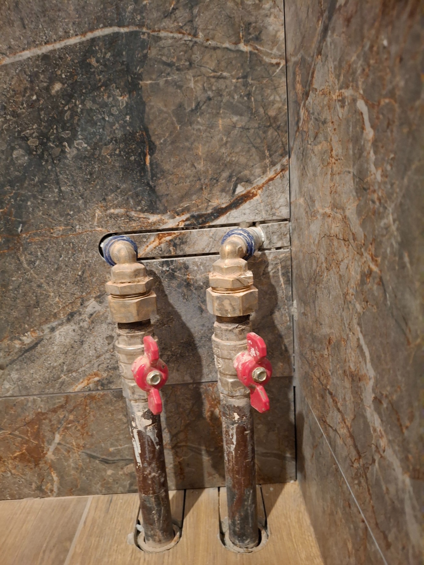 Dwa pionowe rury z zaworami z czerwonymi pokrętłami, wystające z podłogi wyłożonej panelami, na tle ściany z płytek imitujących marmur w odcieniach szarości i brązu.