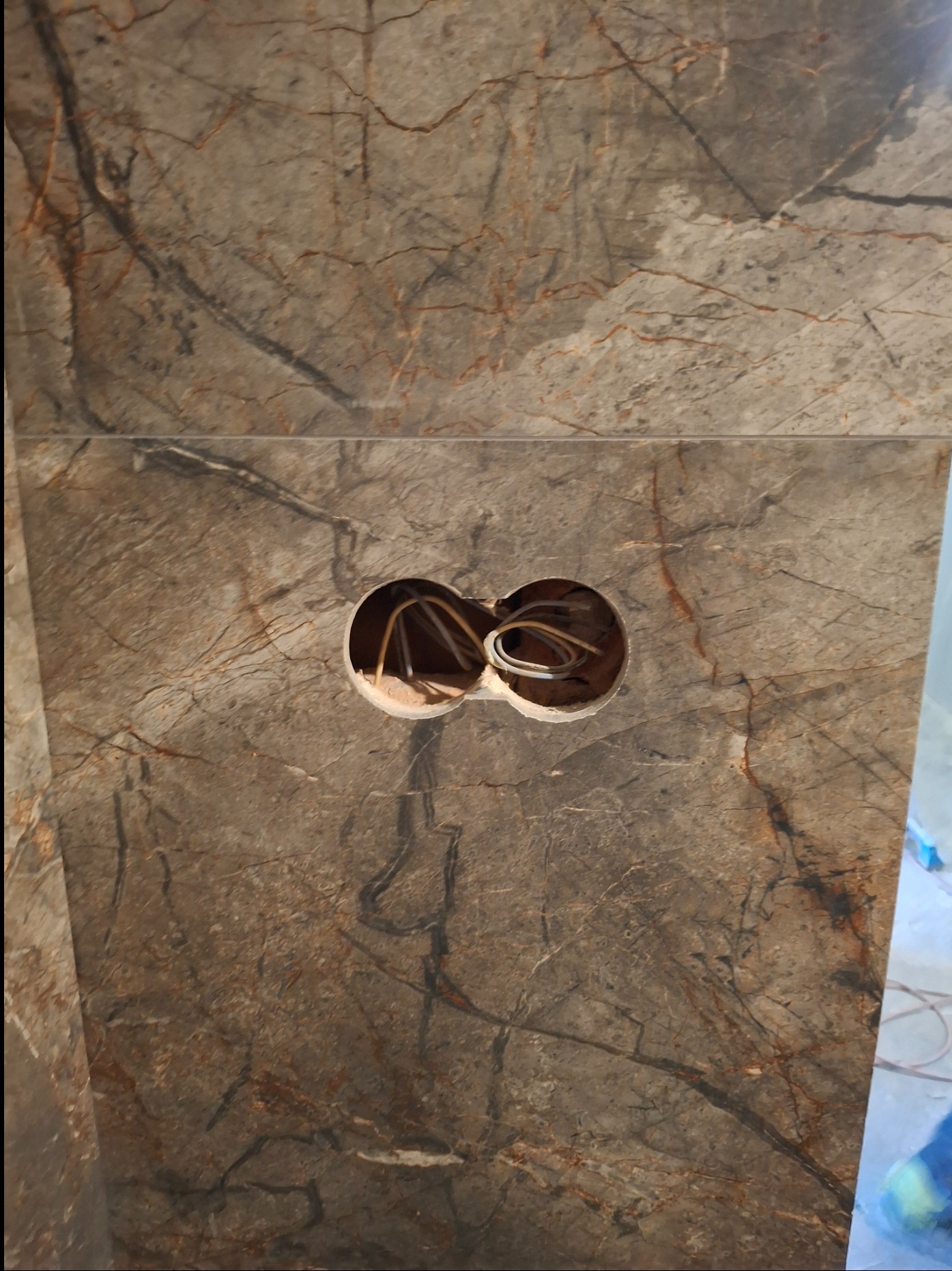 Fragment ściany wyłożonej płytami imitującymi marmur, z dwoma okrągłymi otworami na kable elektryczne.