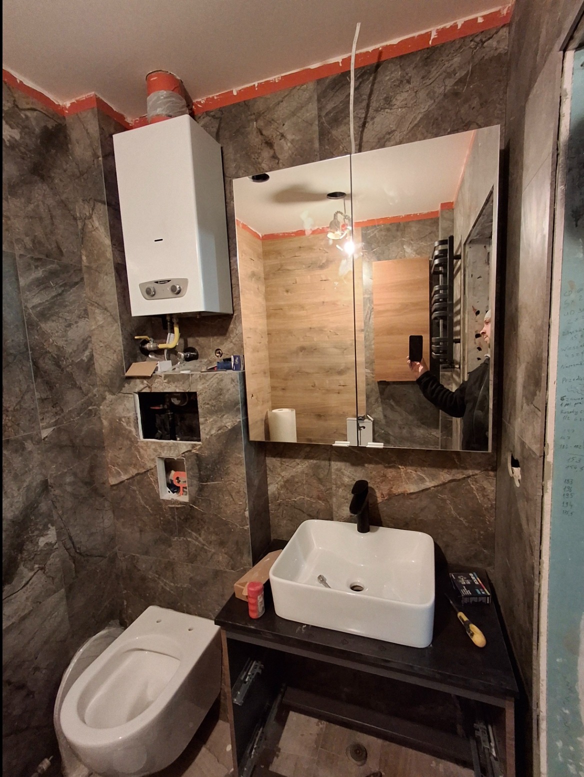 Niedokończona łazienka z marmurowymi płytkami, wiszącą toaletą, białą umywalką na czarnej szafce i lustrem z widocznym odbiciem osoby robiącej zdjęcie, z zamontowanym piecem gazowym...