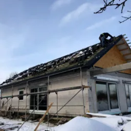 Budowa domu jednorodzinnego w zimowej scenerii: dekarz układa dachówkę na częściowo pokrytym dachu, widoczne rusztowania, ściany z białych pustaków i śnieg na ziemi.