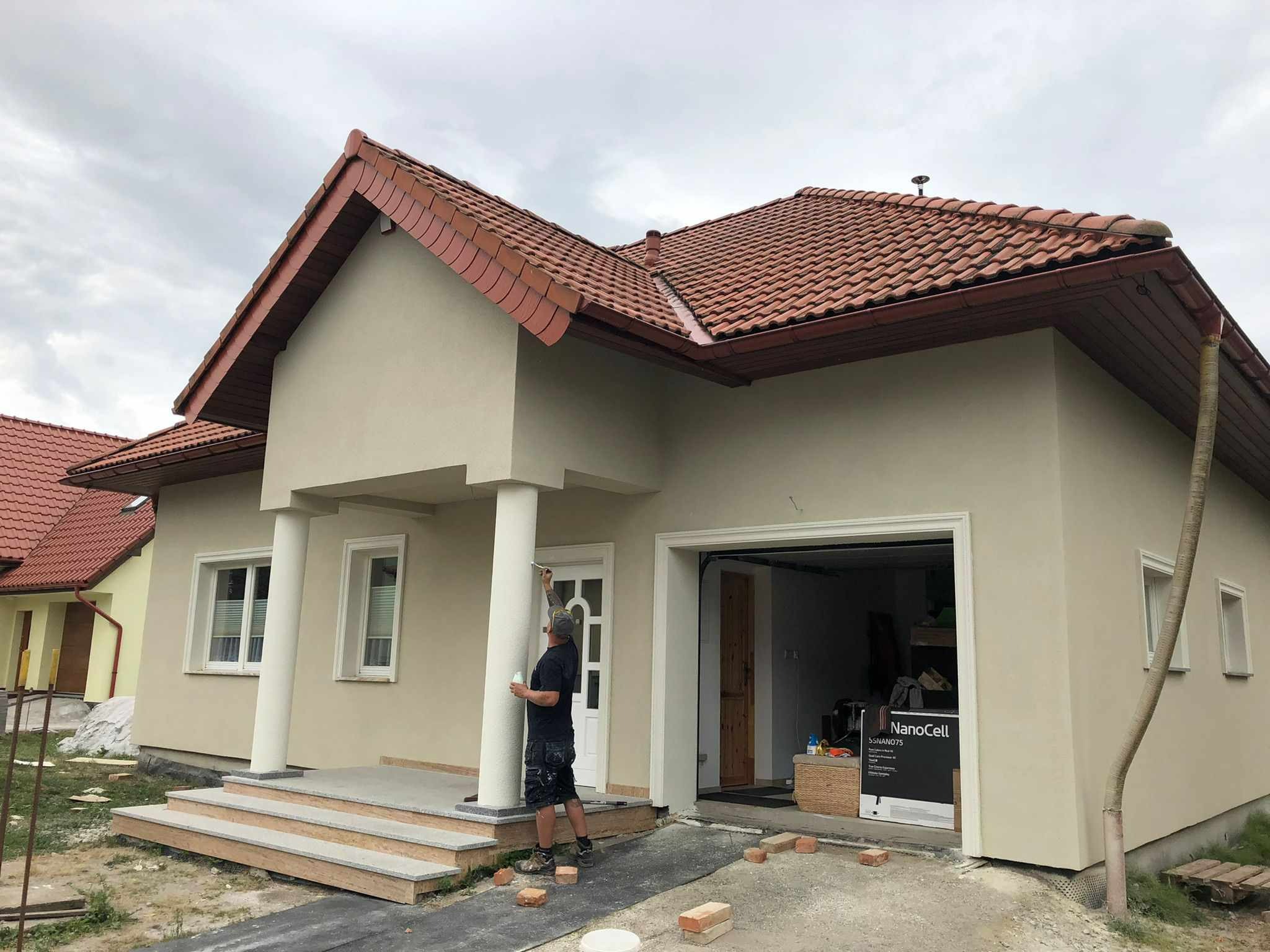 Pracownik maluje kolumnę wejściową domu jednorodzinnego na etapie wykończenia elewacji; widoczna otwarta brama garażowa, czerwona dachówka i częściowo ułożona kostka brukowa.