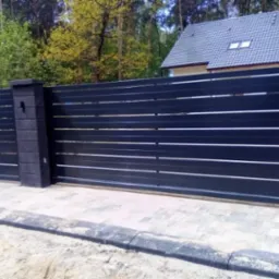 Nowoczesna, przesuwna brama wjazdowa z poziomymi, czarnymi panelami i słupkami z szarej kostki betonowej, widoczna z poziomu chodnika, z domem i drzewami w tle.