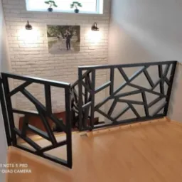 Nowoczesna balustrada schodowa z czarnych, geometrycznych elementów, widok z góry na drewniane schody, ściana z białej cegły dekoracyjnej i oświetlenie punktowe.