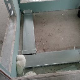 Elektro- Lider usługi elektryczne - Widok z góry na fragment surowego wnętrza w trakcie wykańczania, z widocznymi metalowymi korytkami kablowymi przymocowanymi do podłoża za pomocą białych opasek zaciskowych, oraz pianką montażową...