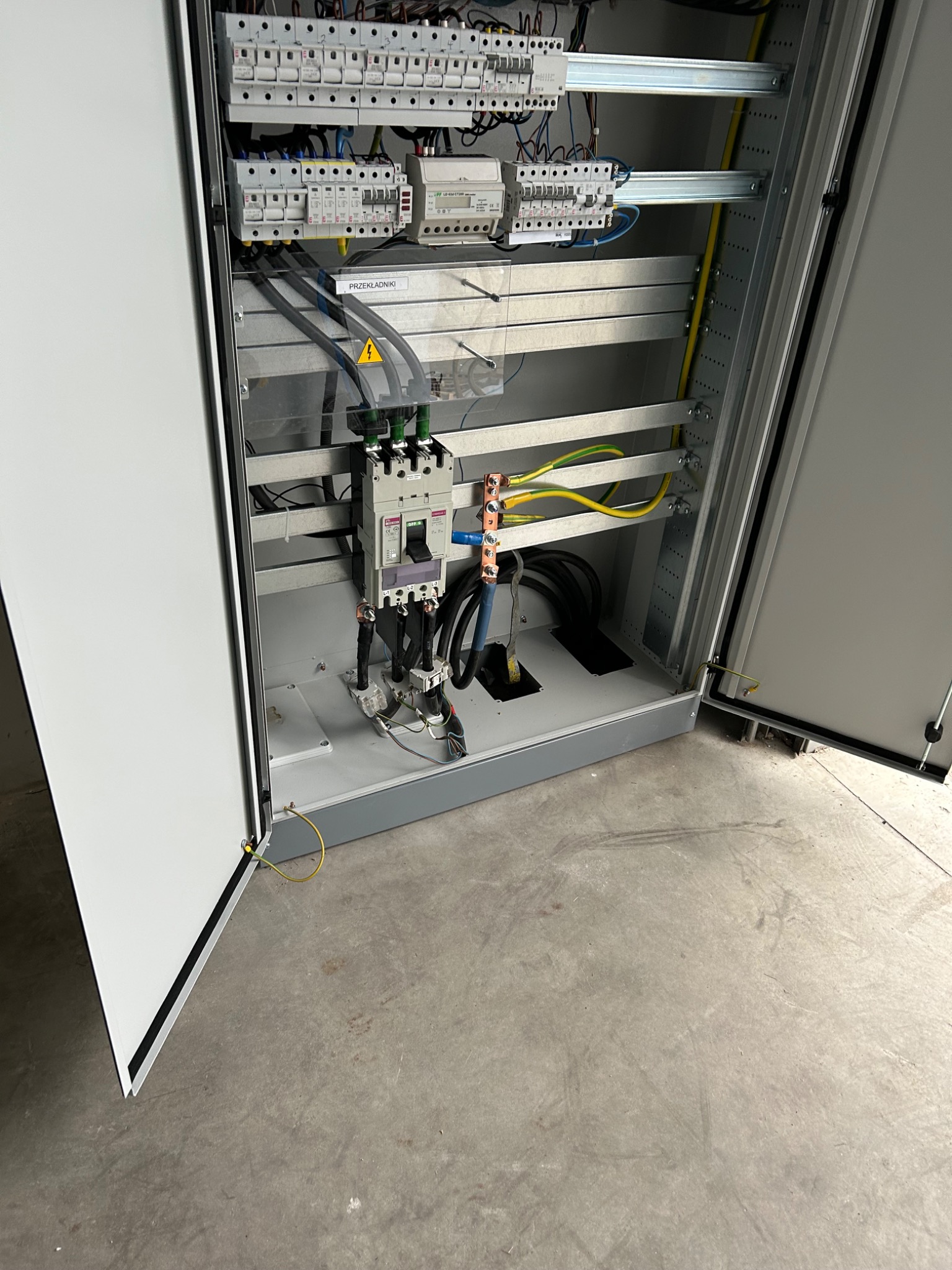 Otwarta szafa elektryczna z widocznymi bezpiecznikami, przewodami w kolorach żółto-zielonym i niebieskim oraz tabliczką ostrzegawczą z symbolem napięcia.