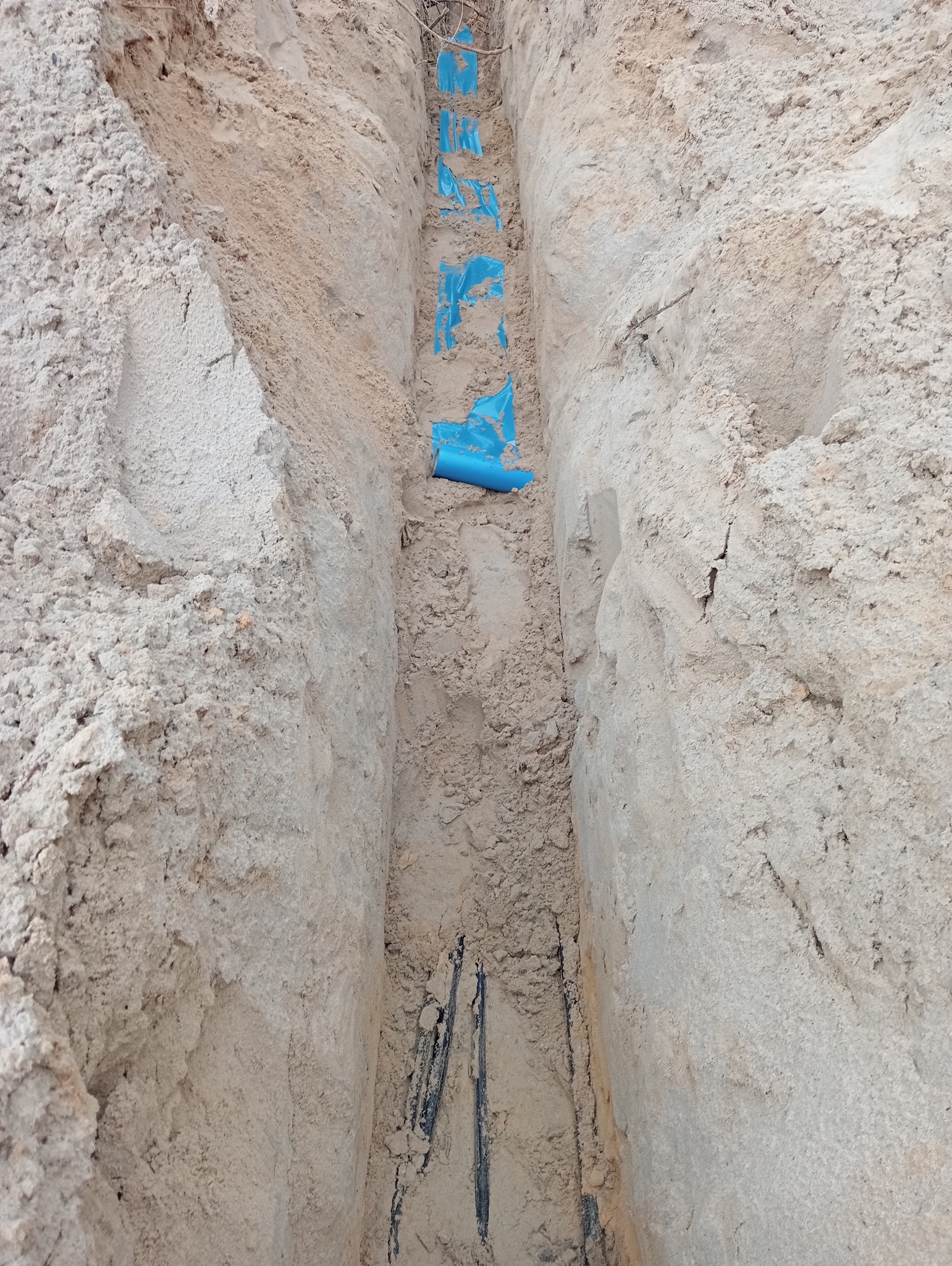 Wykop w piasku z ułożonymi niebieskimi rurami osłonowymi dla kabli elektrycznych, widoczne czarne kable na dnie wykopu.