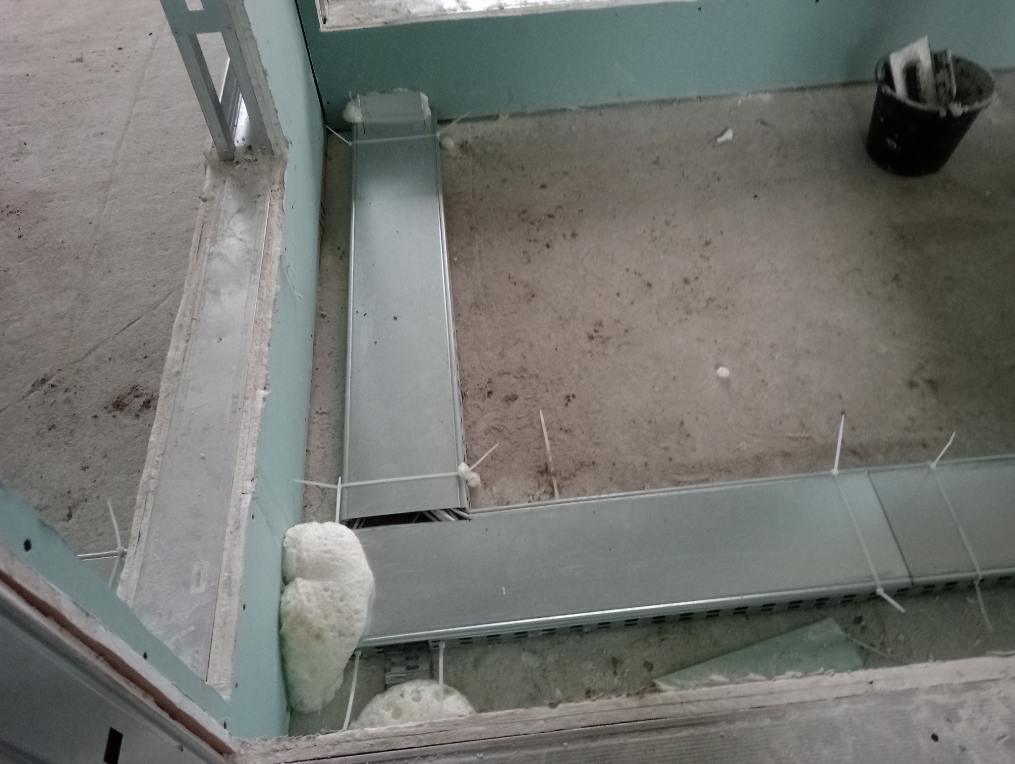 Widok z góry na fragment surowego wnętrza w trakcie wykańczania, z widocznymi metalowymi korytkami kablowymi przymocowanymi do podłoża za pomocą białych opasek zaciskowych, oraz pianką montażową...