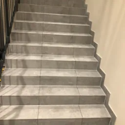 Schody wyłożone szarymi płytkami ceramicznymi z fugami, widok z dołu, czarna metalowa balustrada po lewej stronie, ściana w kolorze ecru po prawej, zielony fragment ściany na górze.