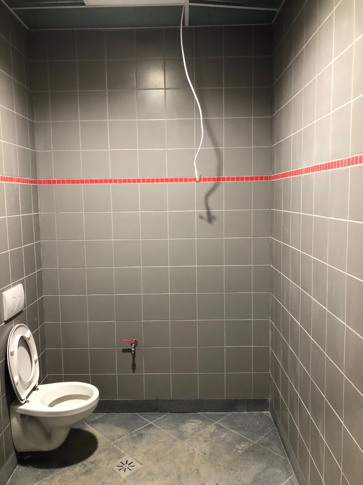 Szara łazienka z białą toaletą, czerwoną listwą dekoracyjną na ścianie i wystającym kablem ze ściany