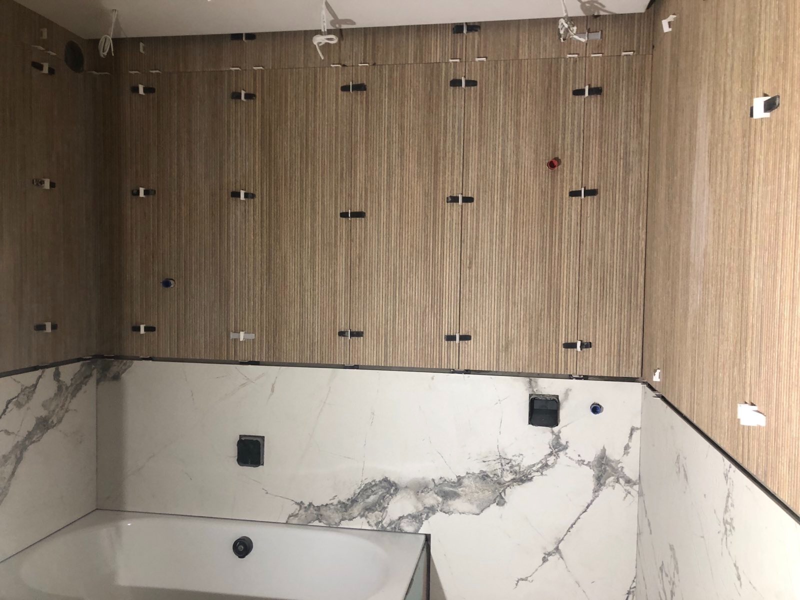 Łazienka w trakcie wykańczania: brązowe płytki imitujące drewno na ścianie, białe marmurowe płytki poniżej, widoczne krzyżyki dystansowe i wystające kable, wanna częściowo widoczna w dolnym rogu.