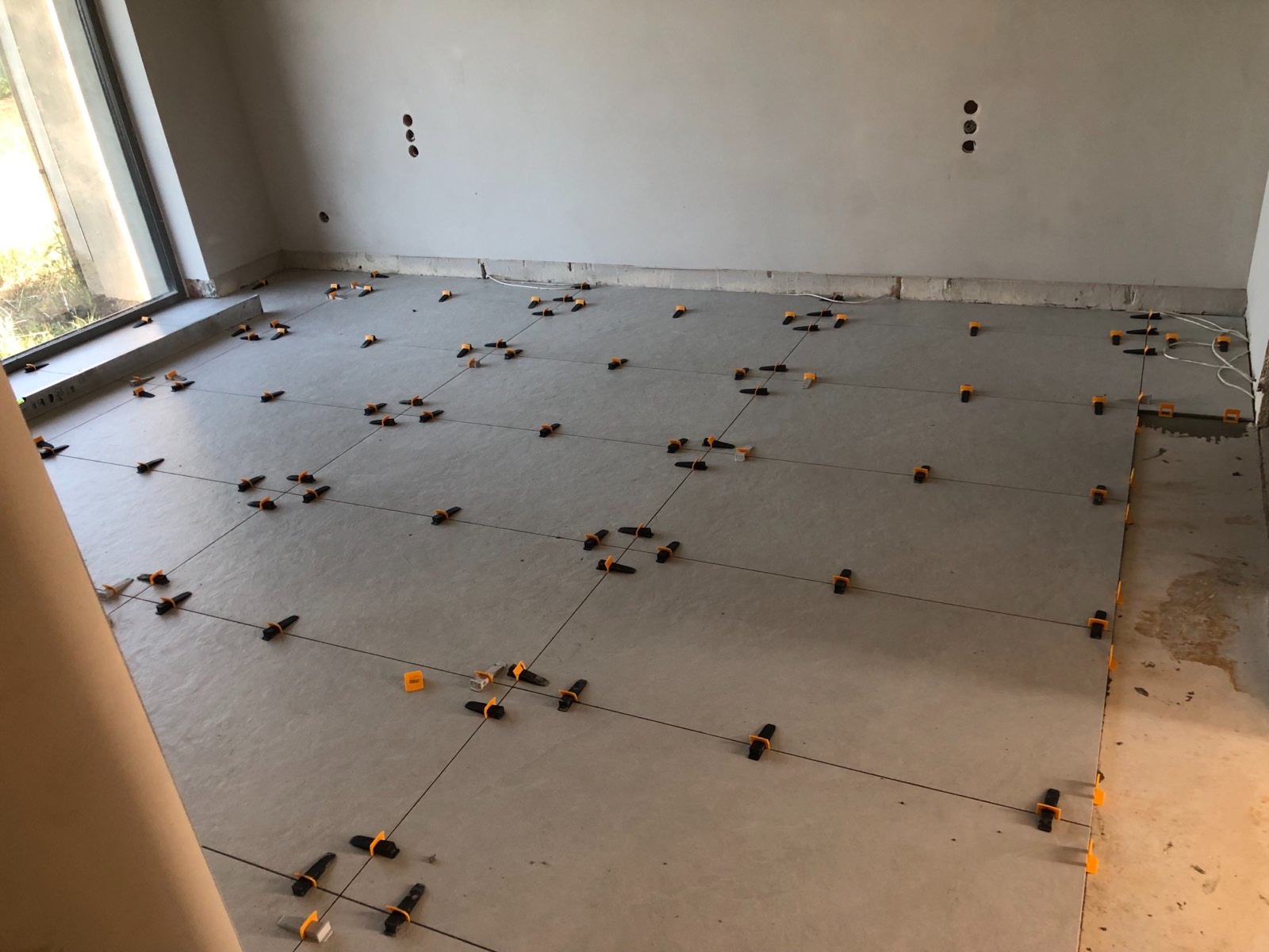 Układanie jasnoszarych płytek podłogowych z użyciem poziomowania w Katowicach, widoczne krzyżyki dystansowe i sznurki wyznaczające linie, ściany w stanie surowym.