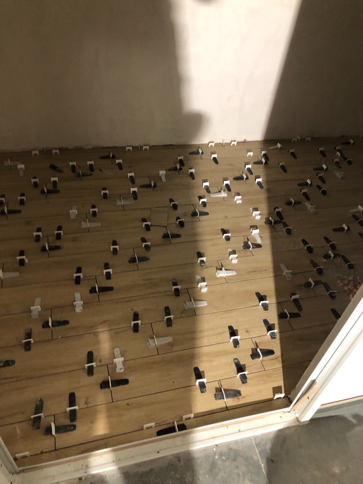 Układanie drewnopodobnych płytek podłogowych z użyciem białych i czarnych klinów poziomujących, widok z góry na fragment podłogi przy ścianie i framudze drzwi.