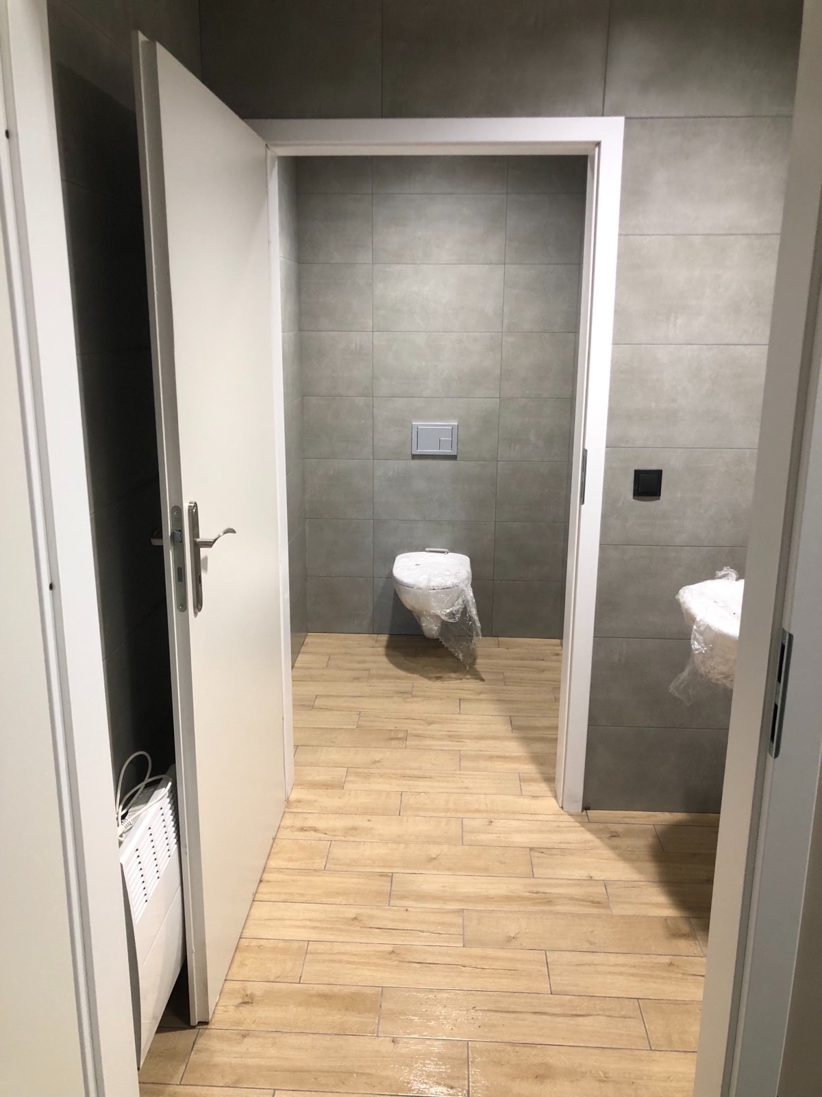 Nowoczesna łazienka w trakcie wykańczania: szare płytki ścienne, drewnopodobne płytki podłogowe, biała armatura zabezpieczona folią, drzwi z widocznym grzejnikiem elektrycznym.