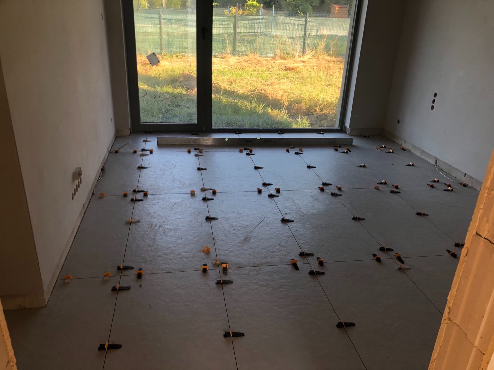 Szara wylewka podłogowa z zamontowanymi elementami poziomującymi i sznurkami krzyżowymi, przygotowana do układania płytek w pomieszczeniu z dużym oknem na zewnątrz.