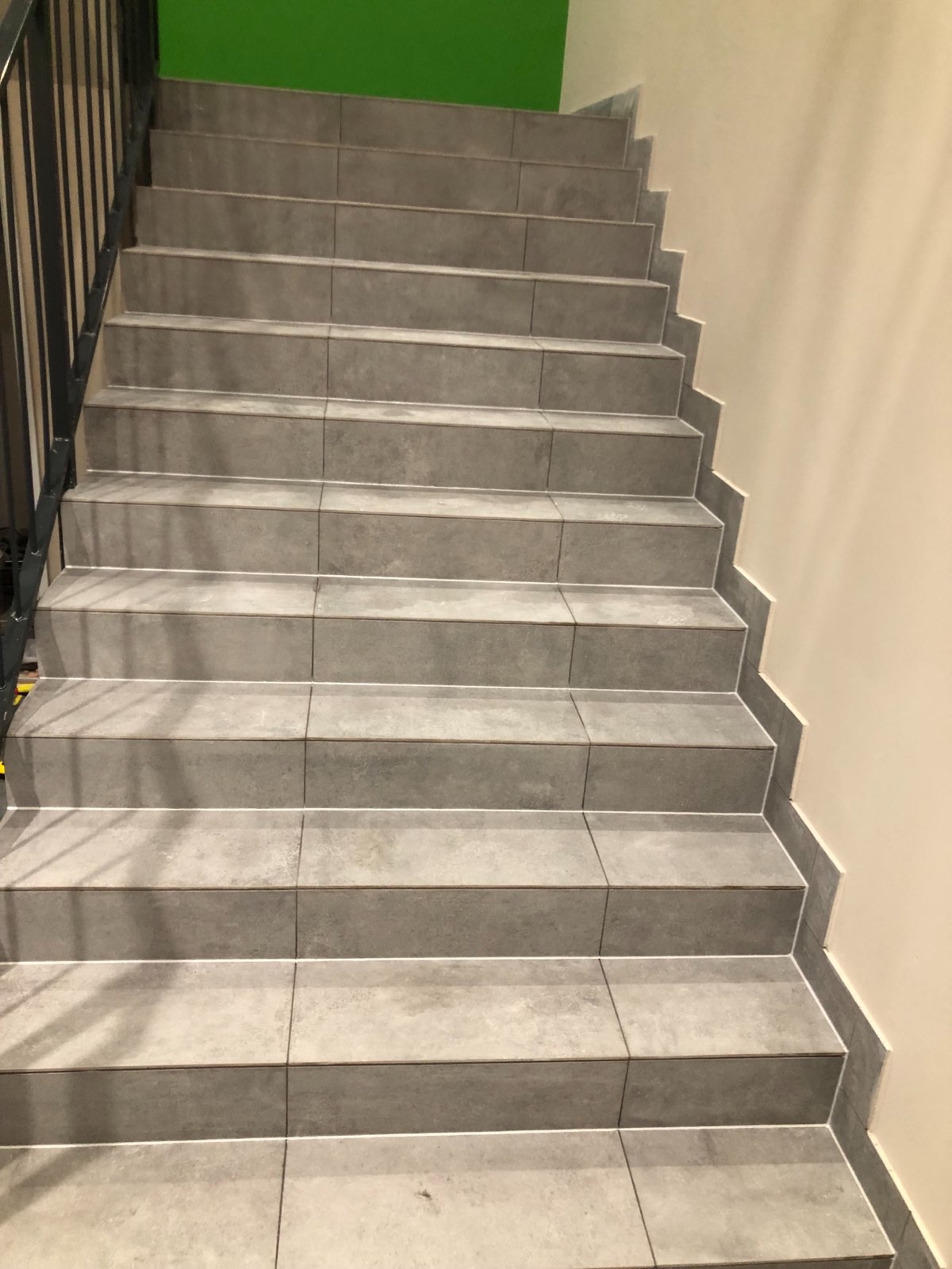 Schody wyłożone szarymi płytkami ceramicznymi z fugami, widok z dołu, czarna metalowa balustrada po lewej stronie, ściana w kolorze ecru po prawej, zielony fragment ściany na górze.
