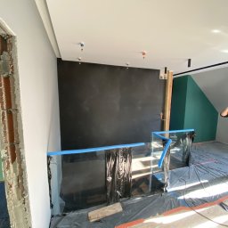 West Bud Ivan Shubak - Pracownik maluje ścianę wałkiem w pomieszczeniu zabezpieczonym folią i taśmą malarską, widoczny fragment surowej ściany po wyburzeniu i instalacje przeciwpożarowe na suficie.