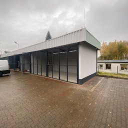 West Bud Ivan Shubak - Zewnętrzny widok nowo wybudowanego pawilonu z szeregiem metalowych, klatkowych boksów, wykończonego jasną elewacją i ciemnym cokołem, na tle pochmurnego nieba i mokrej kostki brukowej.