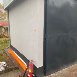 West Bud Ivan Shubak - Świeżo otynkowana ściana budynku z szarą elewacją i czarną, metalową bramą garażową, z widocznym fragmentem rynny, pomarańczowymi zabezpieczeniami i gaśnicą obok.