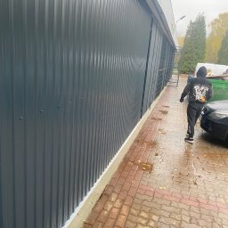West Bud Ivan Shubak - Elewacja budynku przemysłowego z szarej blachy trapezowej, widoczne mocowania śrubowe, obróbka blacharska przy fundamencie, mokry chodnik z kostki brukowej i przechodząca osoba w dresie.