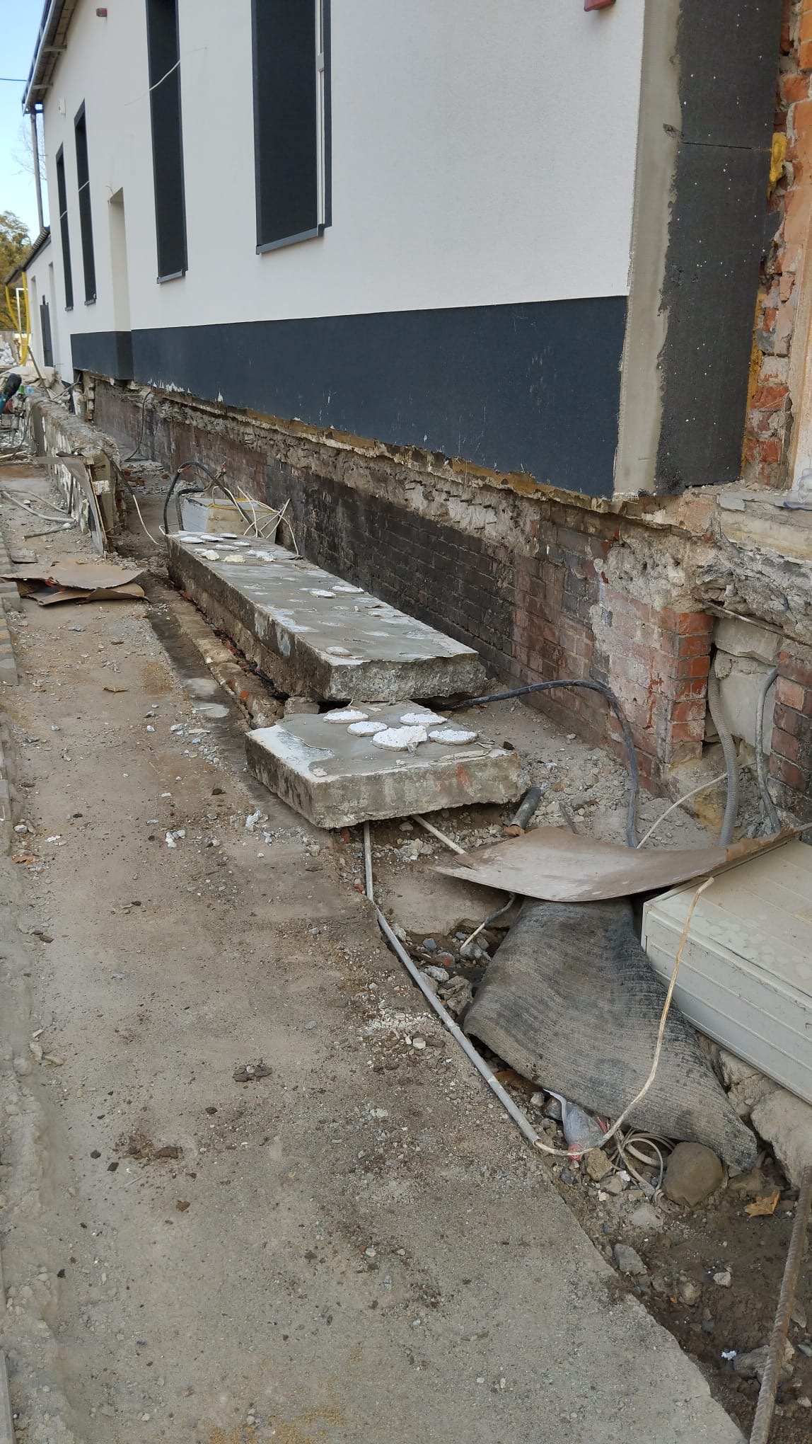 Fragment ściany budynku w trakcie prac, odsłonięte fundamenty i cegły, widoczne przewody i gruz, elementy betonowe na ziemi.