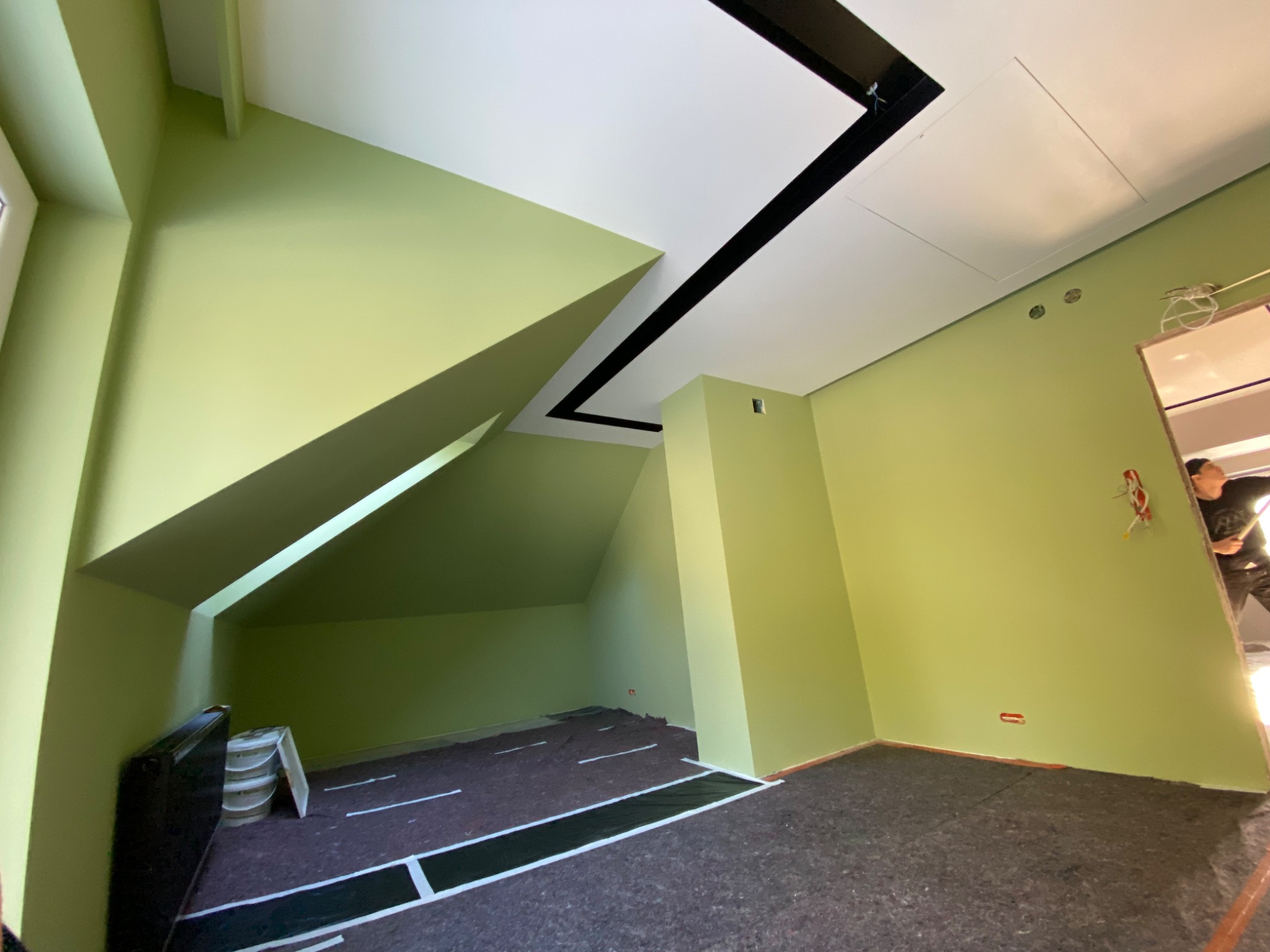 Pomalowane na pistacjowo wnętrze poddasza z nietypowym oknem, zabezpieczona podłoga, widoczny fragment sufitu z czarnymi elementami dekoracyjnymi i pracownik w drzwiach.