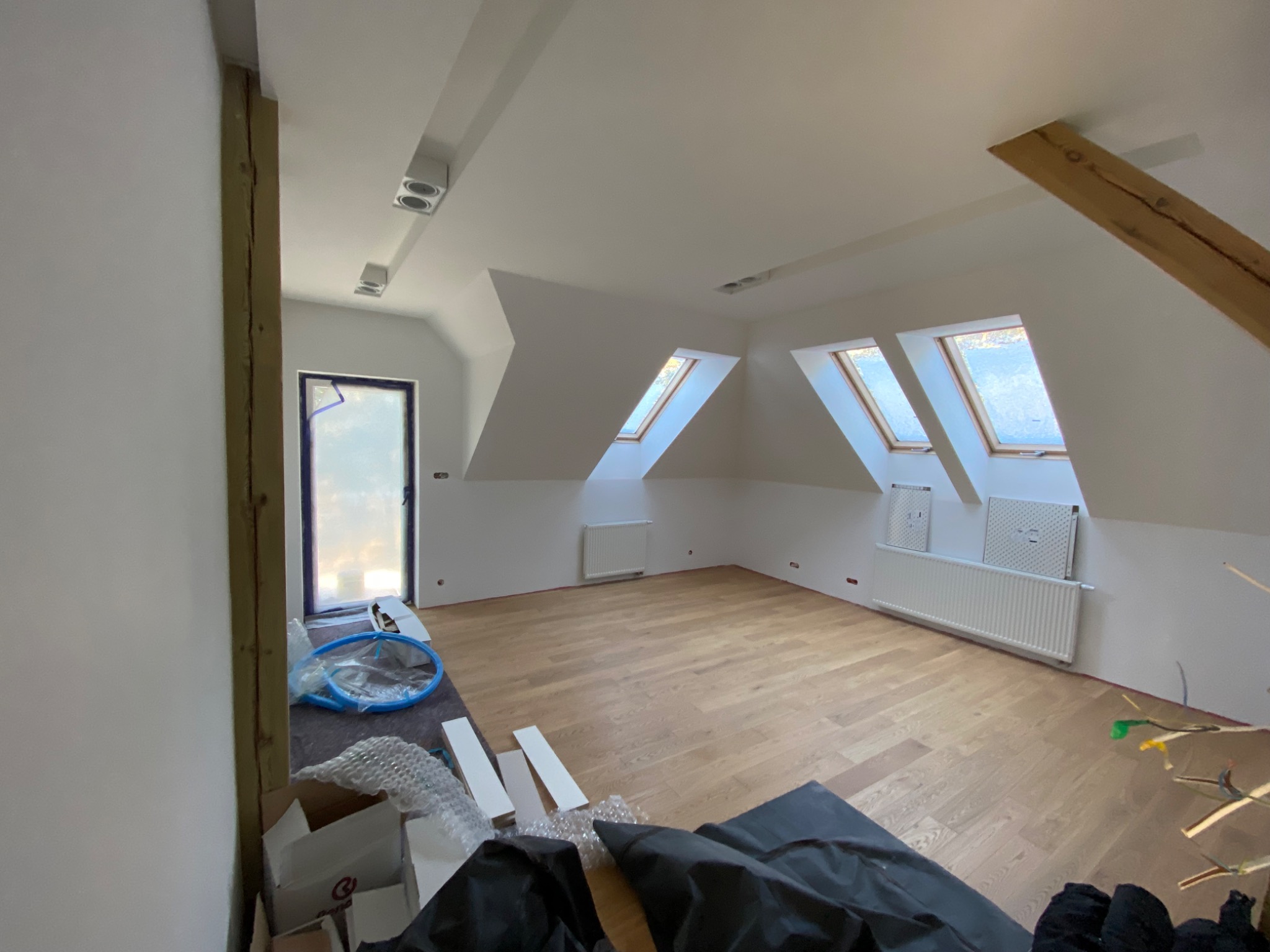 Poddasze po remoncie: drewniana podłoga, okna dachowe, białe ściany z grzejnikami, widoczne elementy konstrukcyjne oraz materiały budowlane w trakcie prac wykończeniowych.