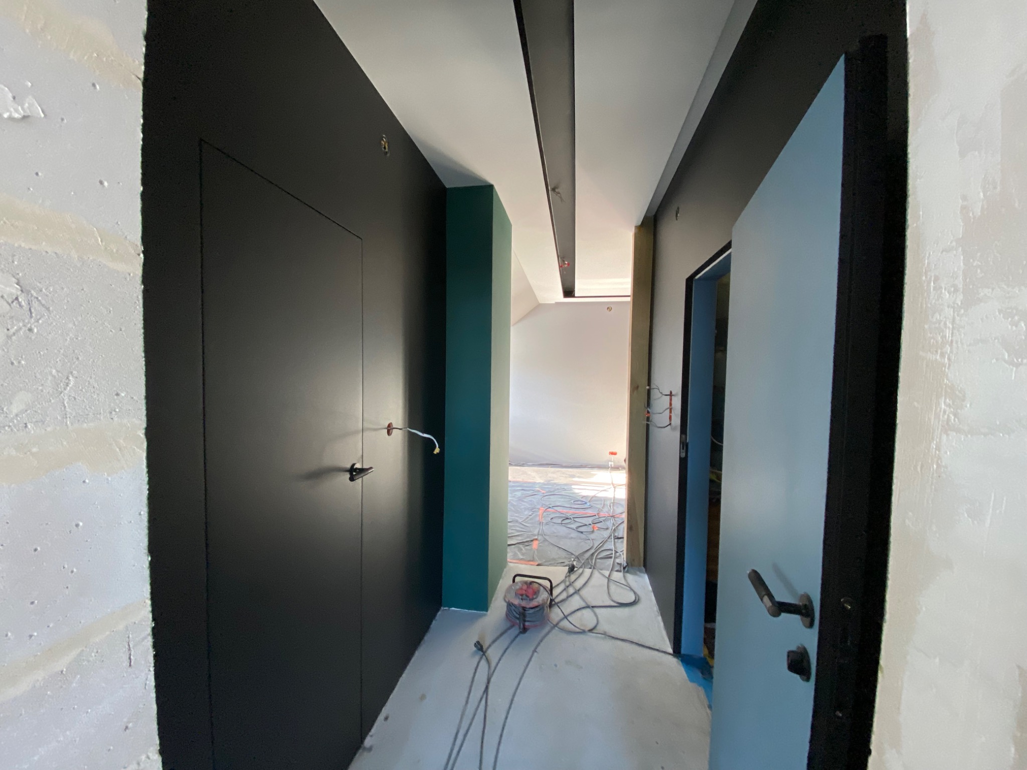 Wnętrze w trakcie remontu: świeżo pomalowane ściany w odcieniach szarości i zieleni, czarne drzwi ukryte, rozciągnięte kable elektryczne i przedłużacz na podłodze, widoczne ślady po gładzi na ścianie.