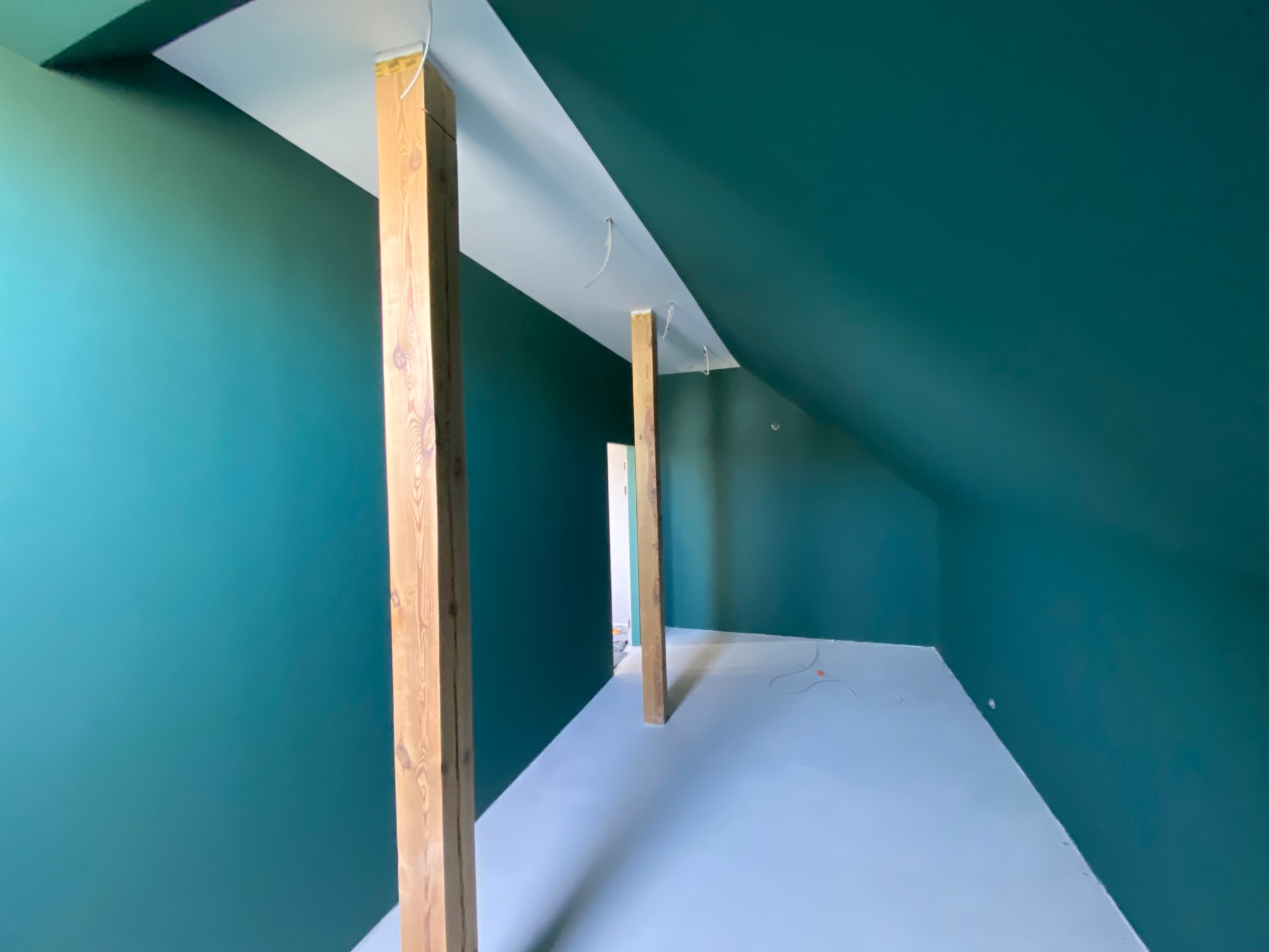 Poddasze po remoncie, ściany pomalowane na ciemny turkus, podłoga jasnoszara, drewniane słupy konstrukcyjne widoczne, wystające kable elektryczne.